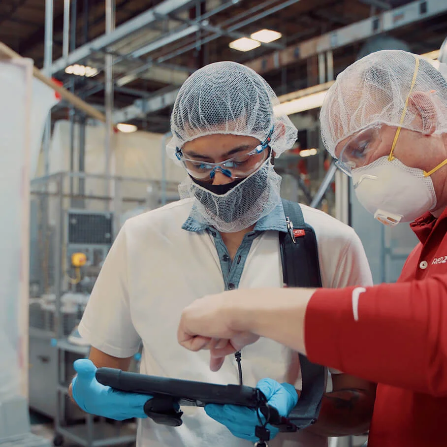 Two factory workers wearing masks, gloves, and hairnets review a tablet in an industrial setting.