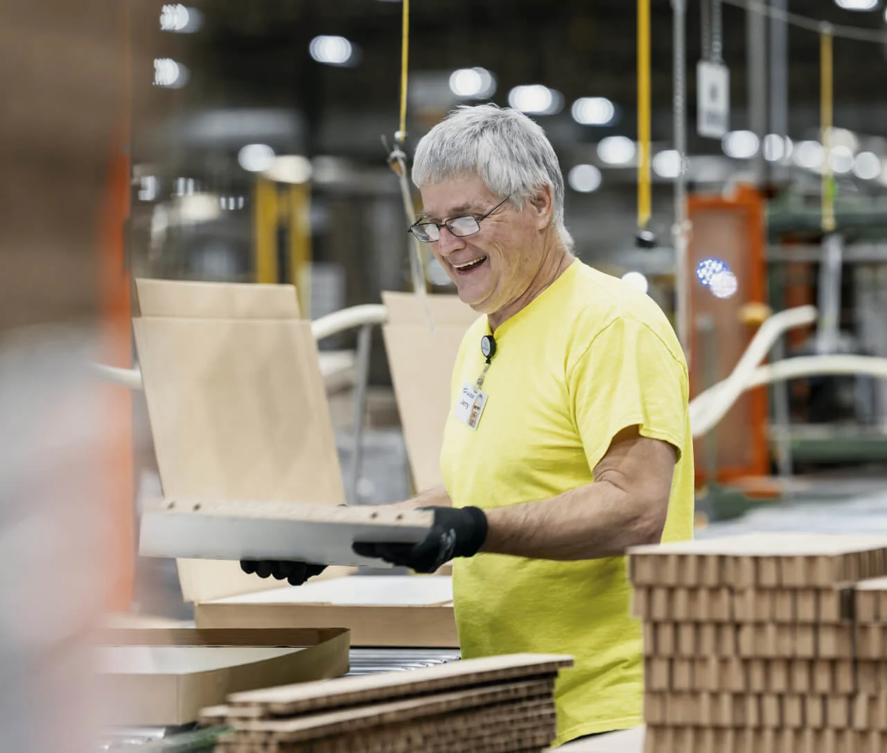 Smiling man in yellow shirt assembling cardboard boxes in a factory setting.