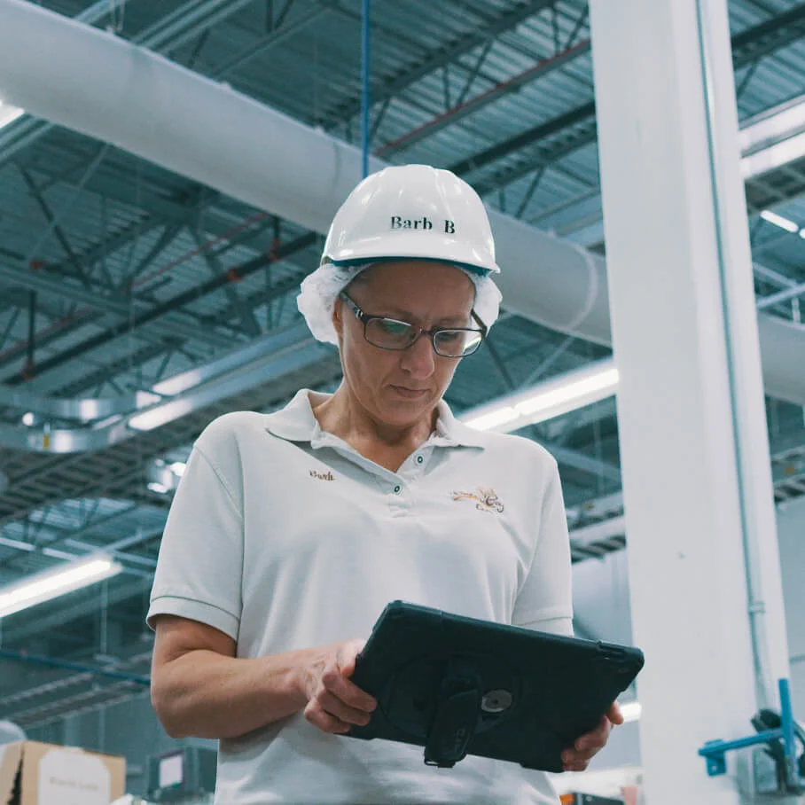 Woman in white safety helmet and glasses using a tablet in an industrial warehouse.