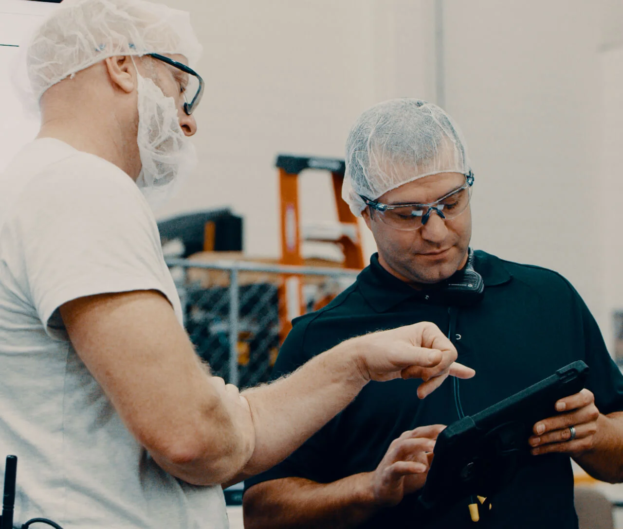 Two factory workers wearing hairnets and safety glasses using a tablet in a warehouse.