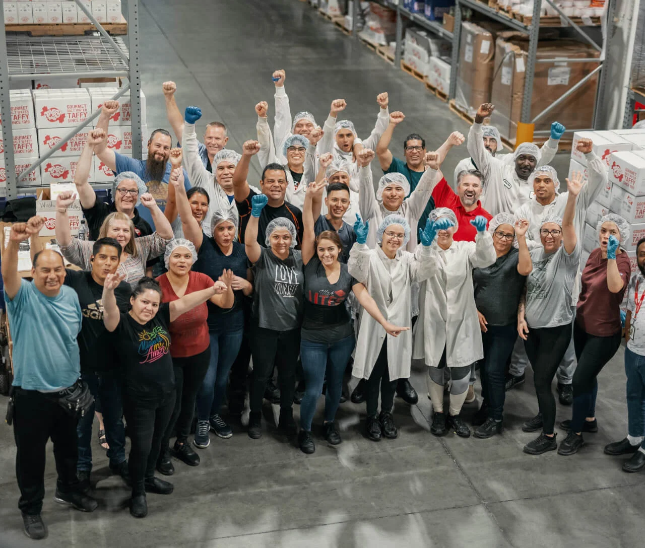 Group of diverse workers in a warehouse raising fists and smiling, some wearing hairnets and gloves.