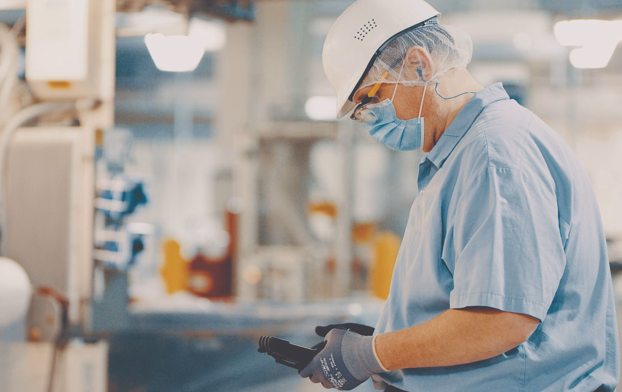 Factory worker wearing mask, gloves, and helmet inspecting a black item in an industrial setting.