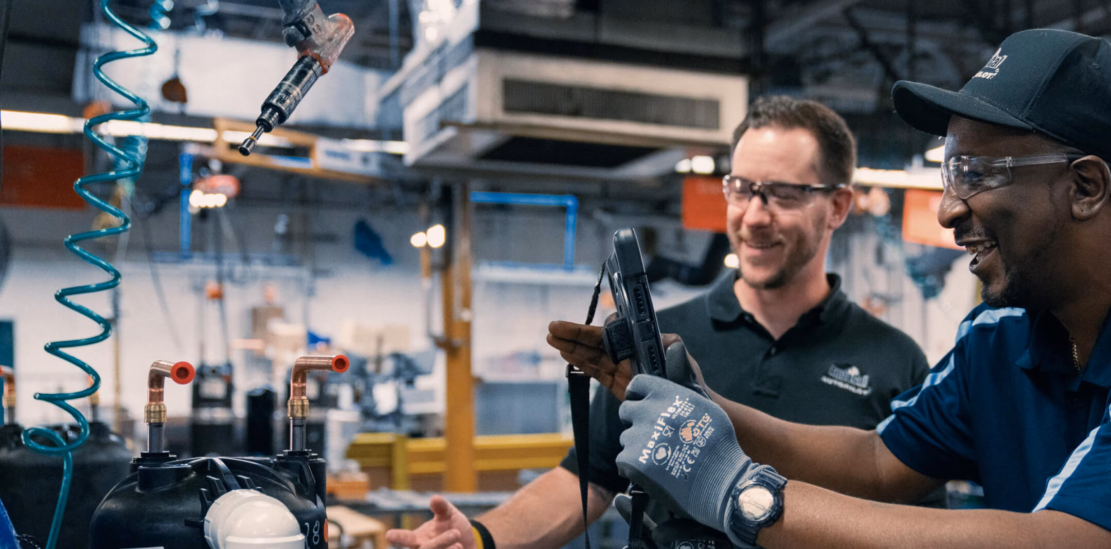 Two factory workers wearing safety glasses and gloves operate machinery controls inside an industrial workshop.