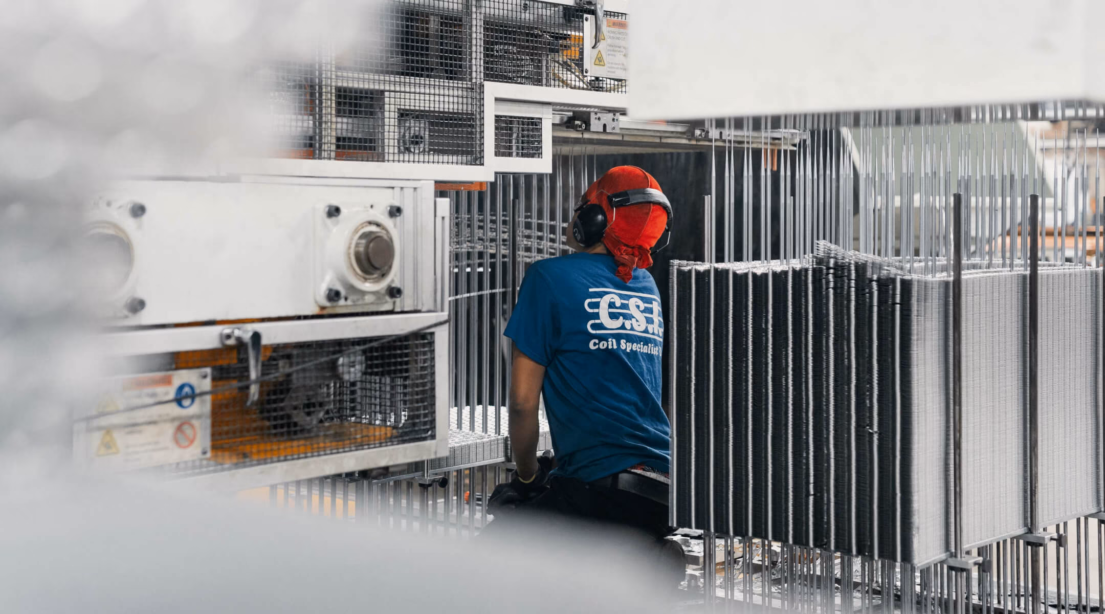 Worker wearing a blue shirt with 'Coil Specialist' text, red head covering, and ear protection operating industrial machinery with metal coils.