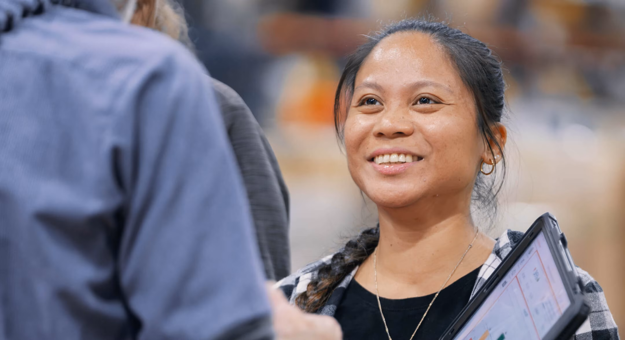 Smiling woman with braided hair holding a tablet, talking to another person whose back is to the camera.