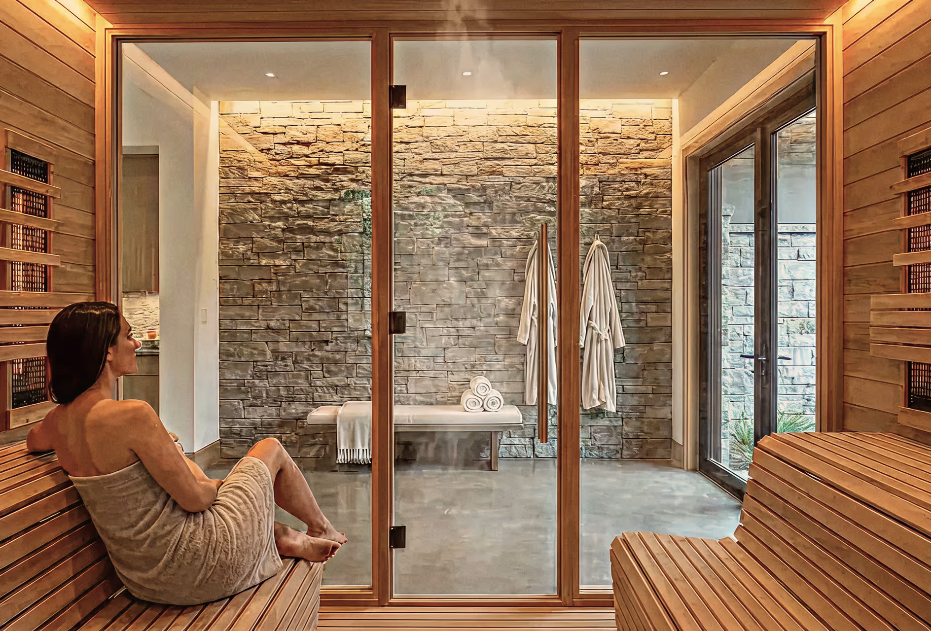 Woman wrapped in towel sitting inside a wooden sauna with glass doors showing a steam room with stone walls, two robes, and a bench with rolled towels.