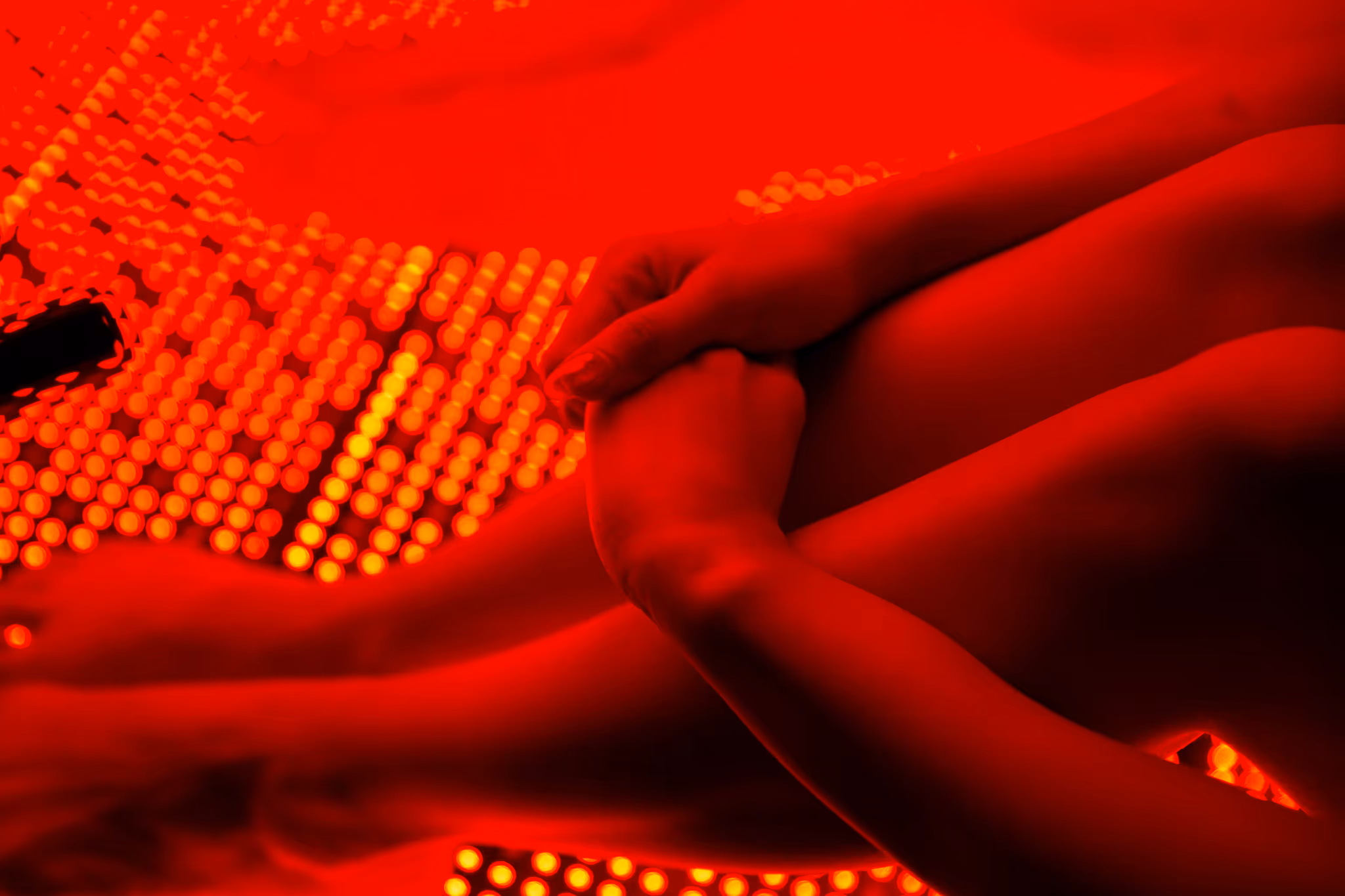 Person wrapped in a white towel sitting in a wooden sauna with red light therapy panel on the wall.