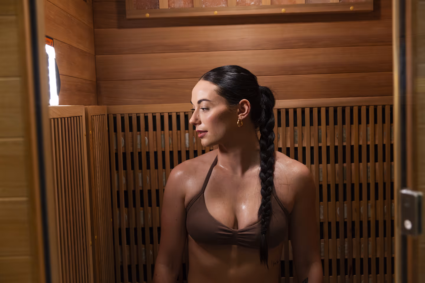 Woman with a long braid sitting inside a wooden infrared sauna, looking to the side.