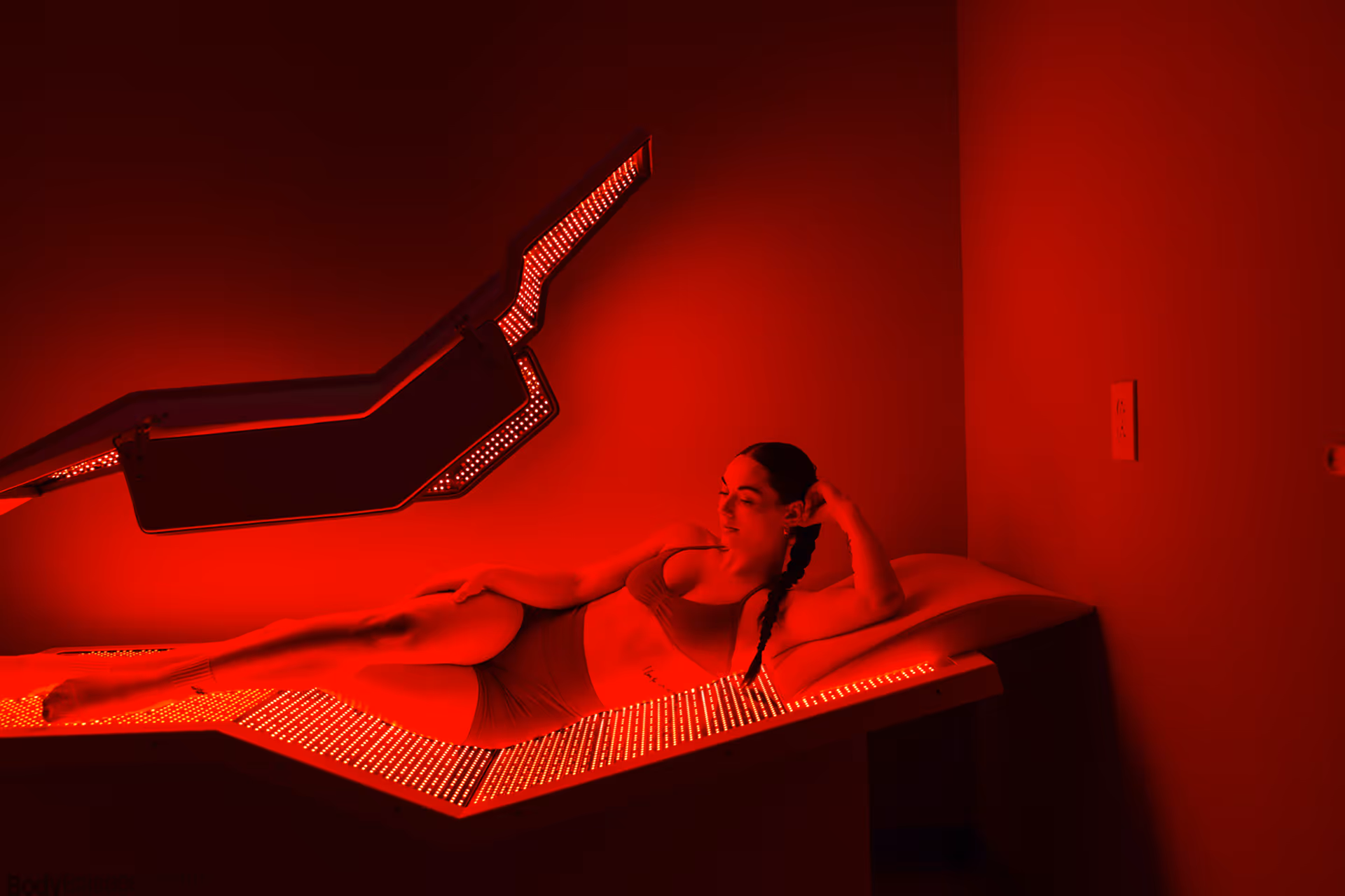 Woman in sportswear lying on a red LED therapy bed in a dimly lit room.