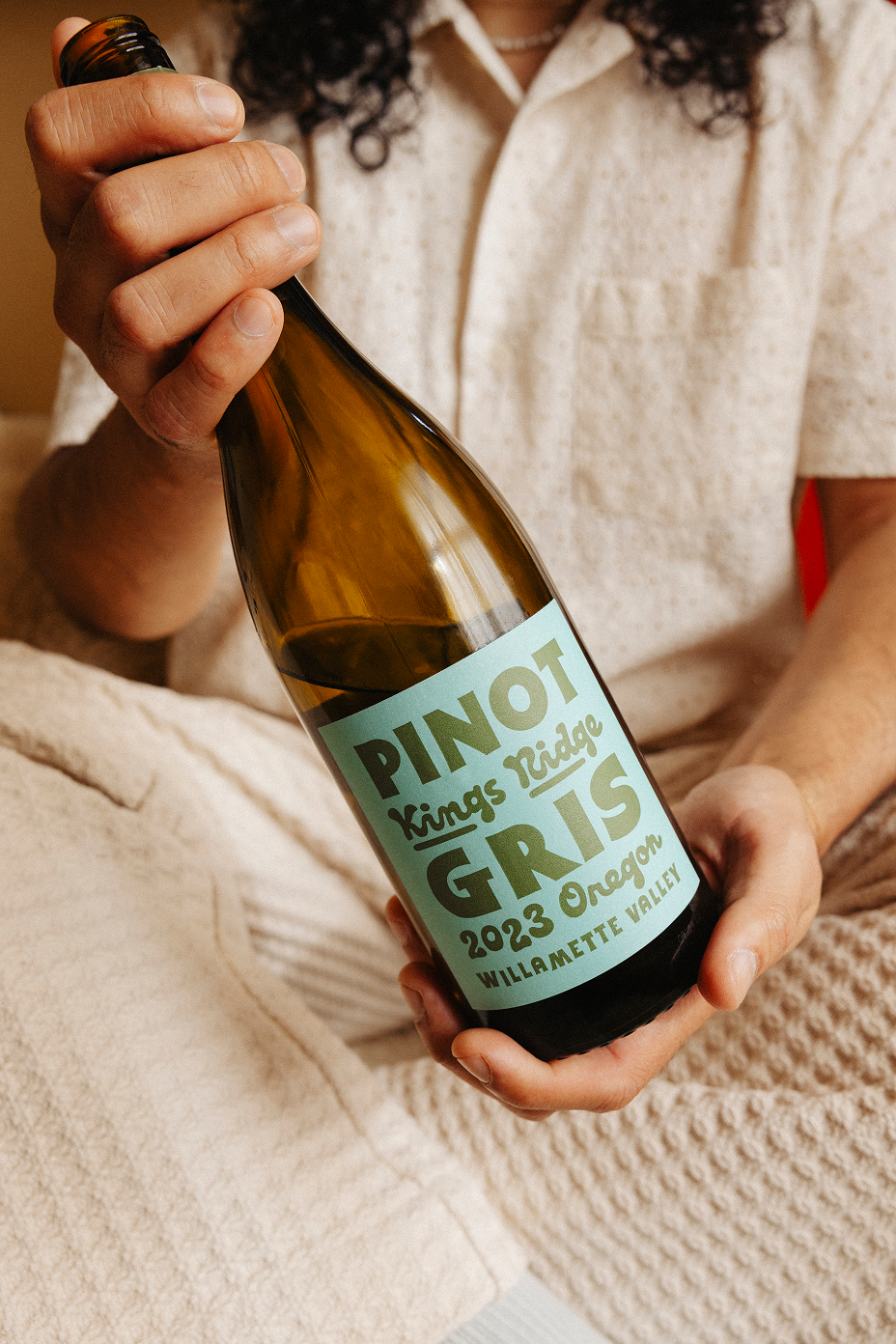 Person wearing a light beige outfit holding a bottle of Pinot Gris wine labeled Kings Ridge 2023 Oregon Willamette Valley.