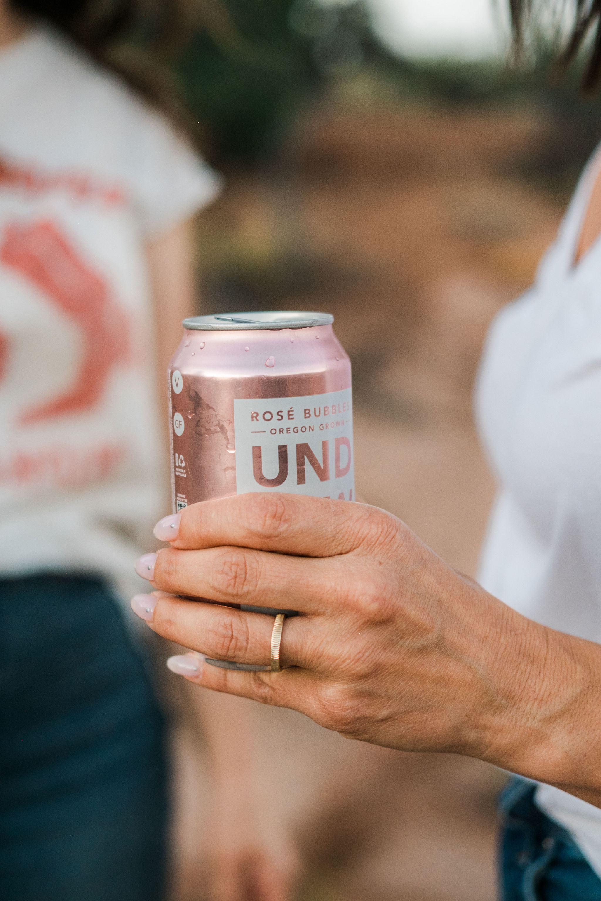 Hand with a gold ring holding a pink can of rosé bubbles drink outdoors.