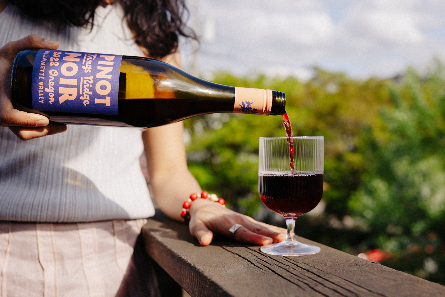 Person pouring red Pinot Noir wine from a bottle into a glass outdoors on a sunny day.