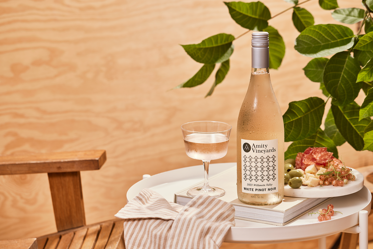 Bottle of Amity Vineyards White Pinot Noir wine and a glass on a table with a charcuterie plate, striped cloth, and green leaves in the background.