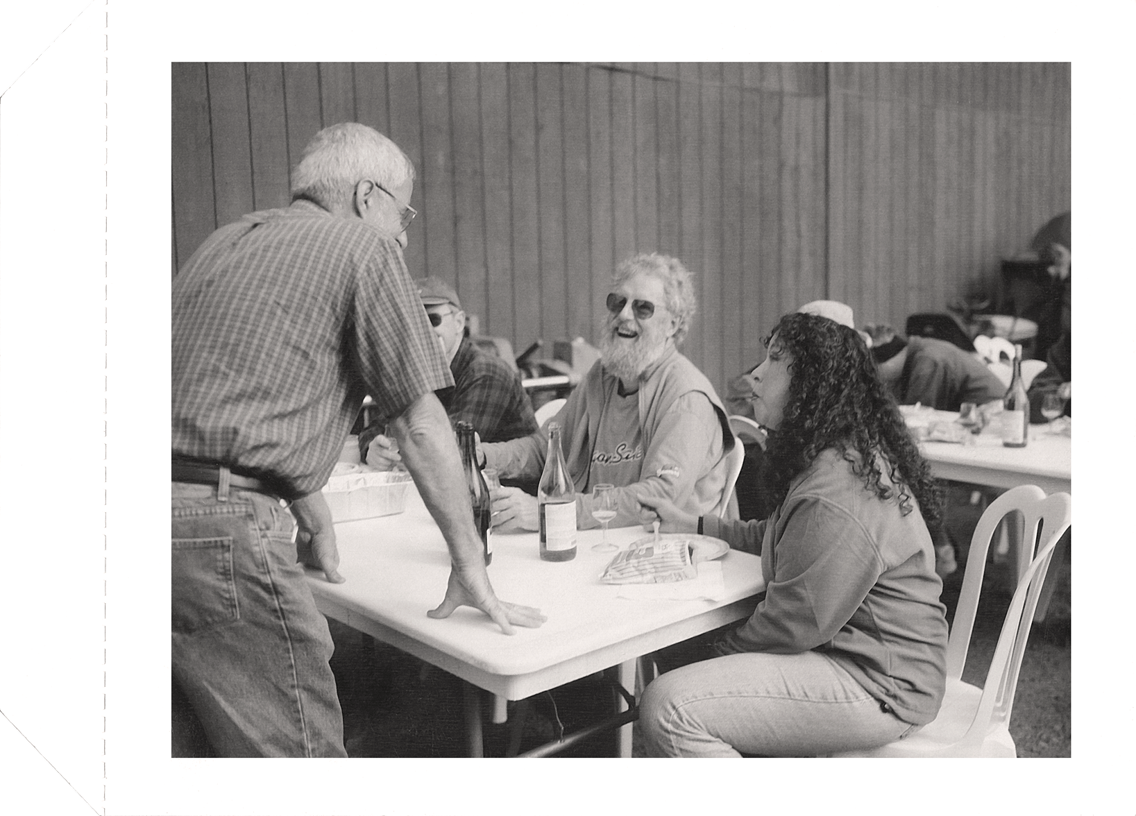Four people gathered around a table engaged in conversation with wine bottles and glasses, two men seated and one woman seated, while an older man leans on the table.