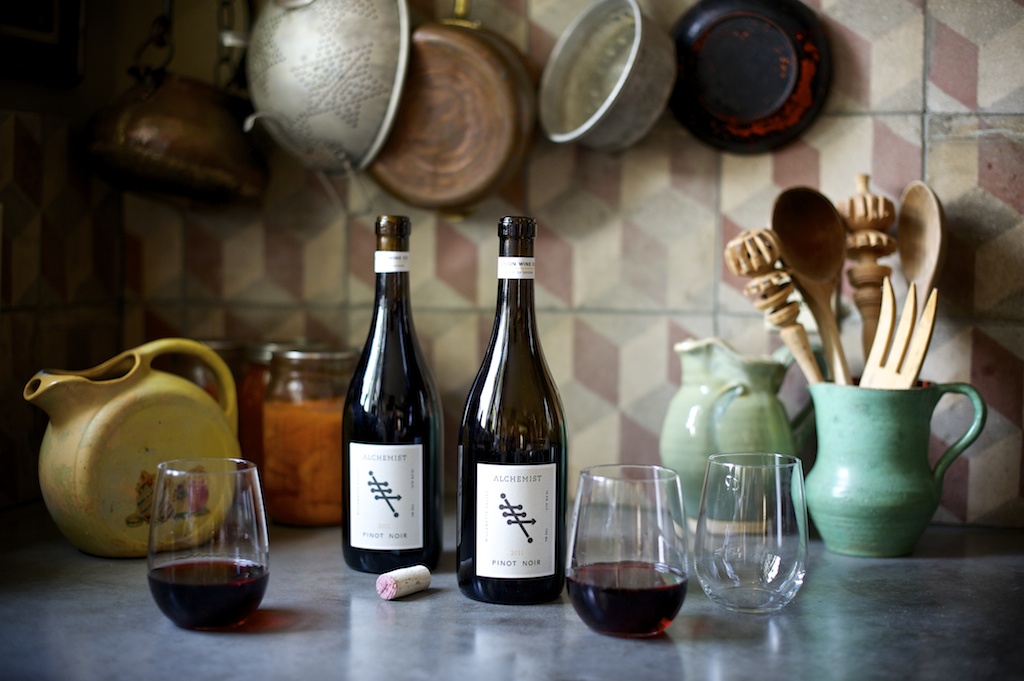 Two bottles of Alchemist Pinot Noir wine with one cork and three drinking glasses, two filled with red wine, on a kitchen counter with vintage kitchenware in the background.