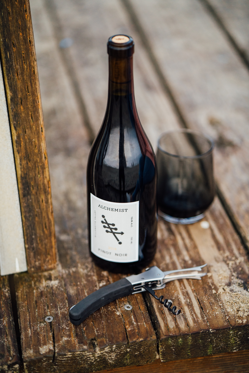 Bottle of Alchemist Pinot Noir wine, a glass with dark liquid, and a corkscrew on a weathered wooden surface.