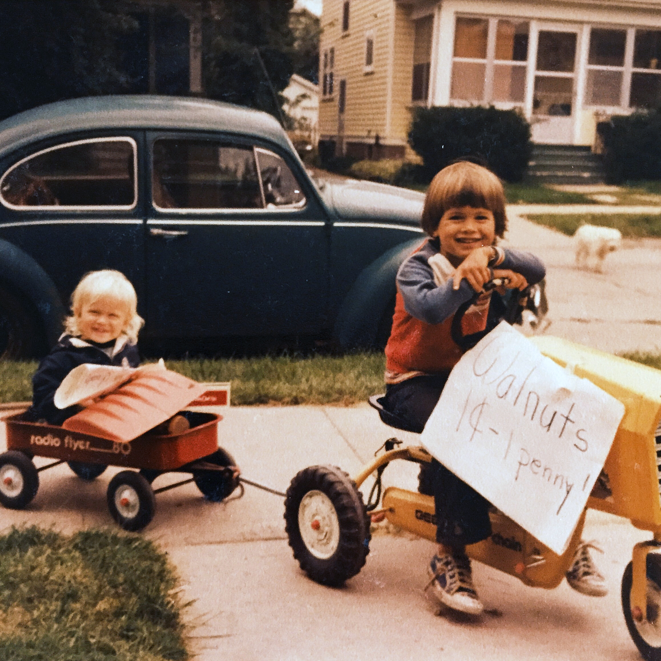 Two children smiling outdoors; one rides a yellow pedal tractor with a sign reading 'Walnuts 14-1 penny!' while pulling a red wagon with the other child sitting inside.