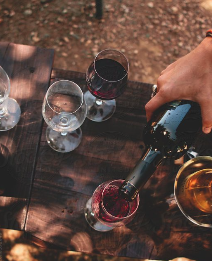 Hand pouring red wine from a bottle into a glass on a wooden table with other empty and filled glasses.