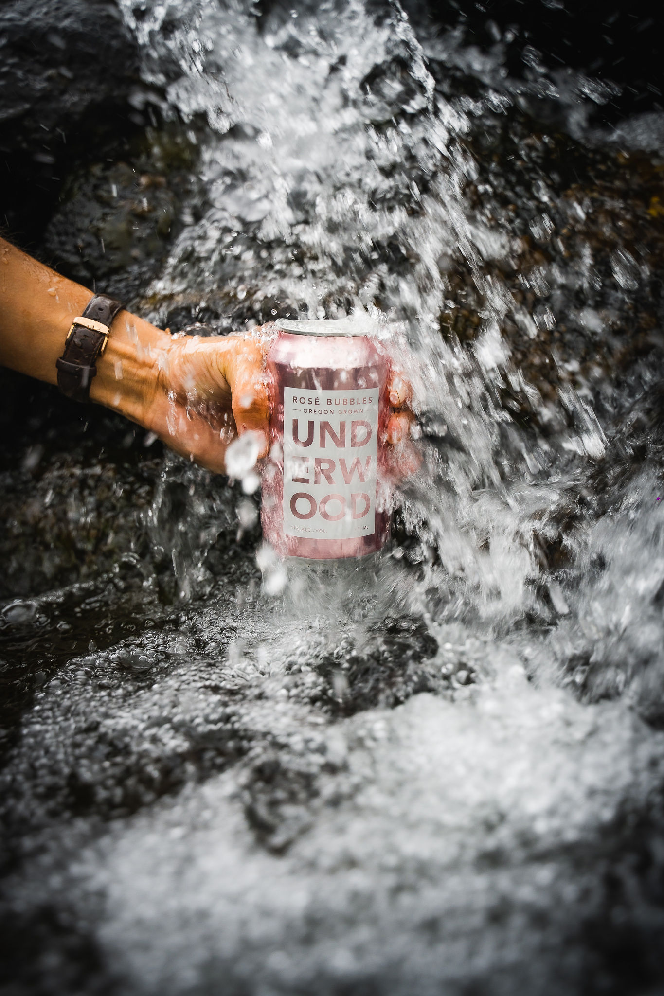 Hand holding a can of Underwood Rosé Bubbles under flowing water in a natural setting.