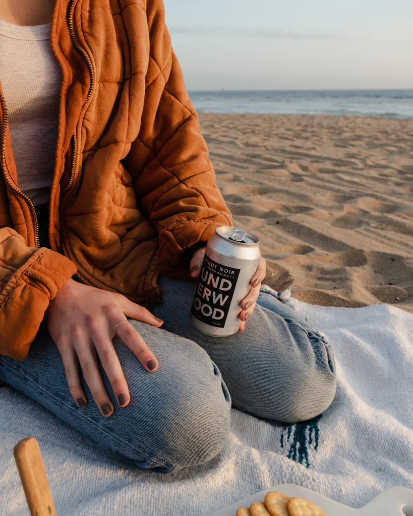 Person sitting on a blanket at the beach wearing a rust-colored jacket and holding a can of Underwood Pinot Noir wine.
