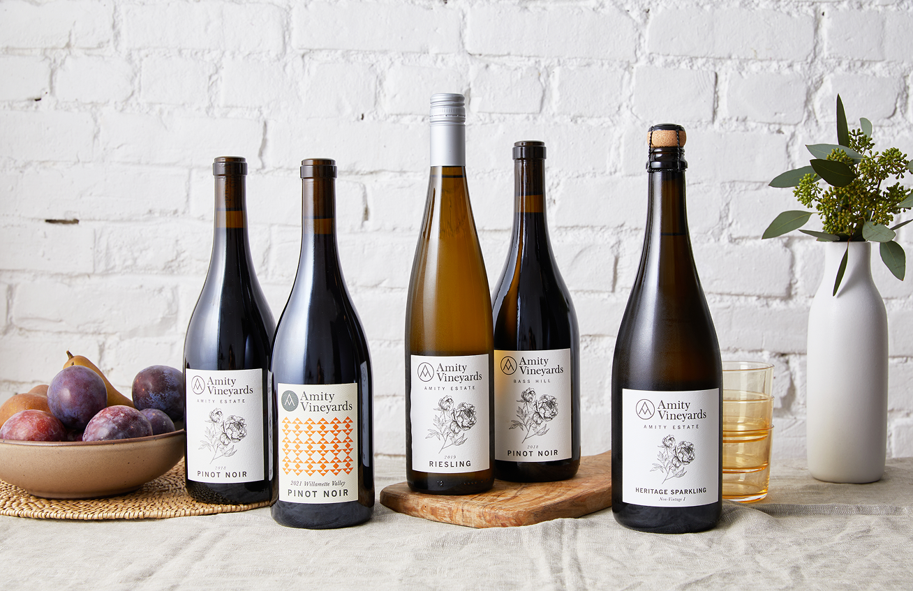 Five bottles of Amity Vineyards wine including Pinot Noir, Riesling, and Heritage Sparkling arranged on a table with a bowl of plums and a white vase with greenery.