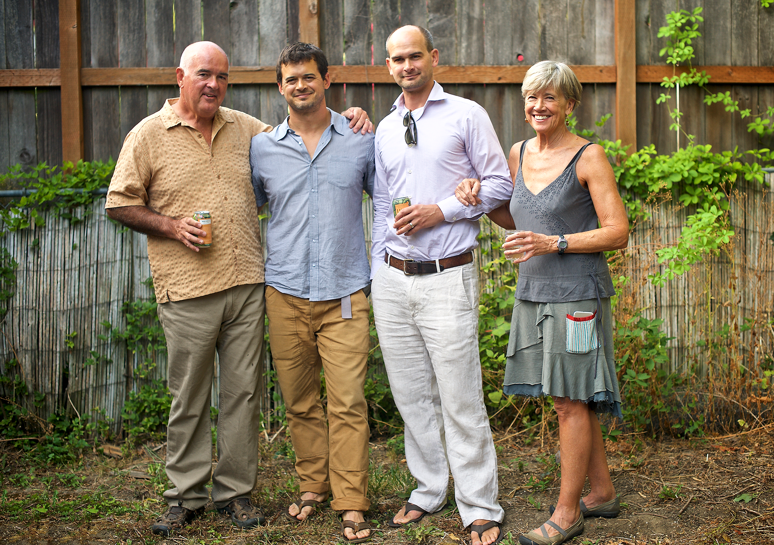 Four adults standing side by side outdoors near a wooden fence, smiling, two holding cans and one holding a glass.