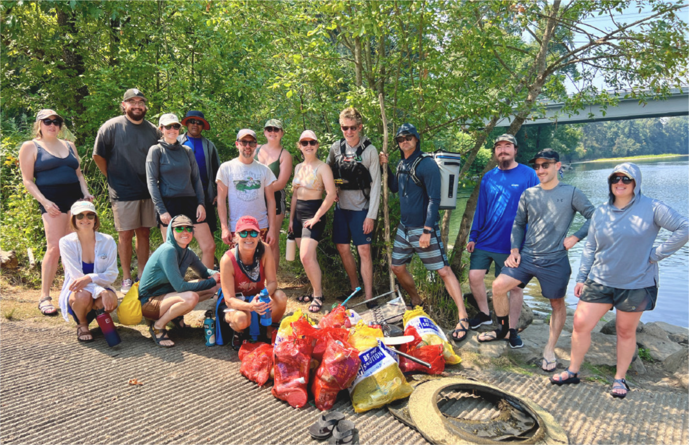 Group of people posing outdoors by a river with collected bags of trash, indicating a community cleanup event.