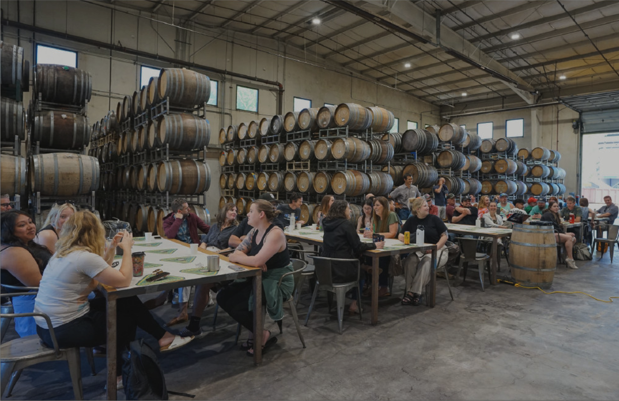 Group of people sitting at long tables inside a warehouse with stacked wooden barrels along the walls.