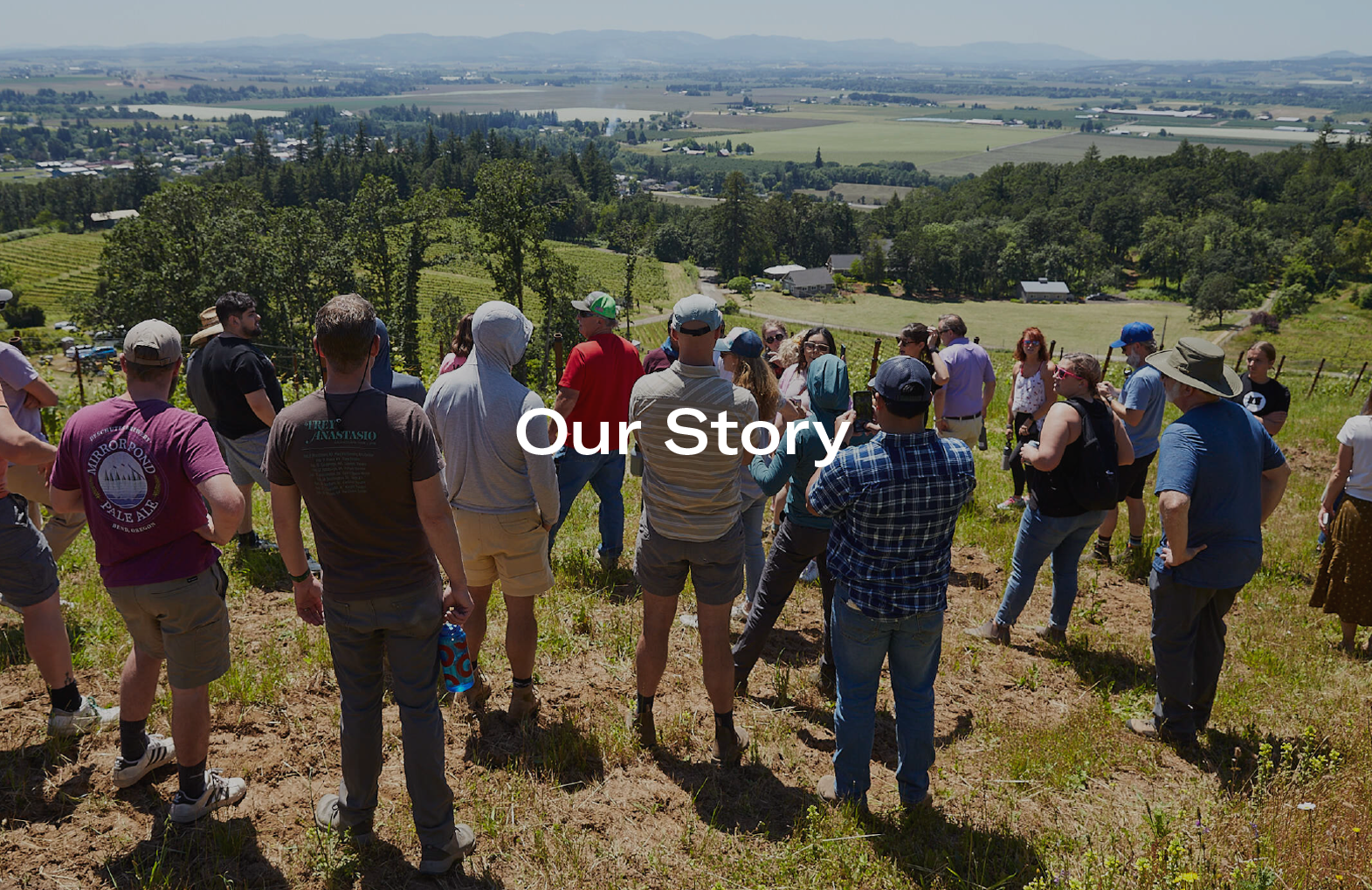 Group of people gathered on a hillside overlooking a rural landscape with fields and forests under a clear sky.