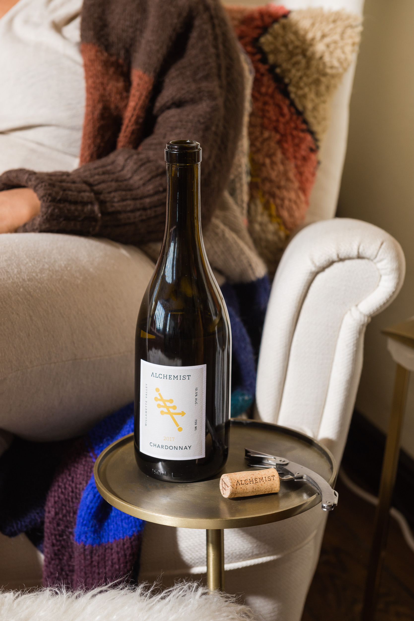 Bottle of 2017 Alchemist Chardonnay wine on a brass side table with a cork and corkscrew beside it.