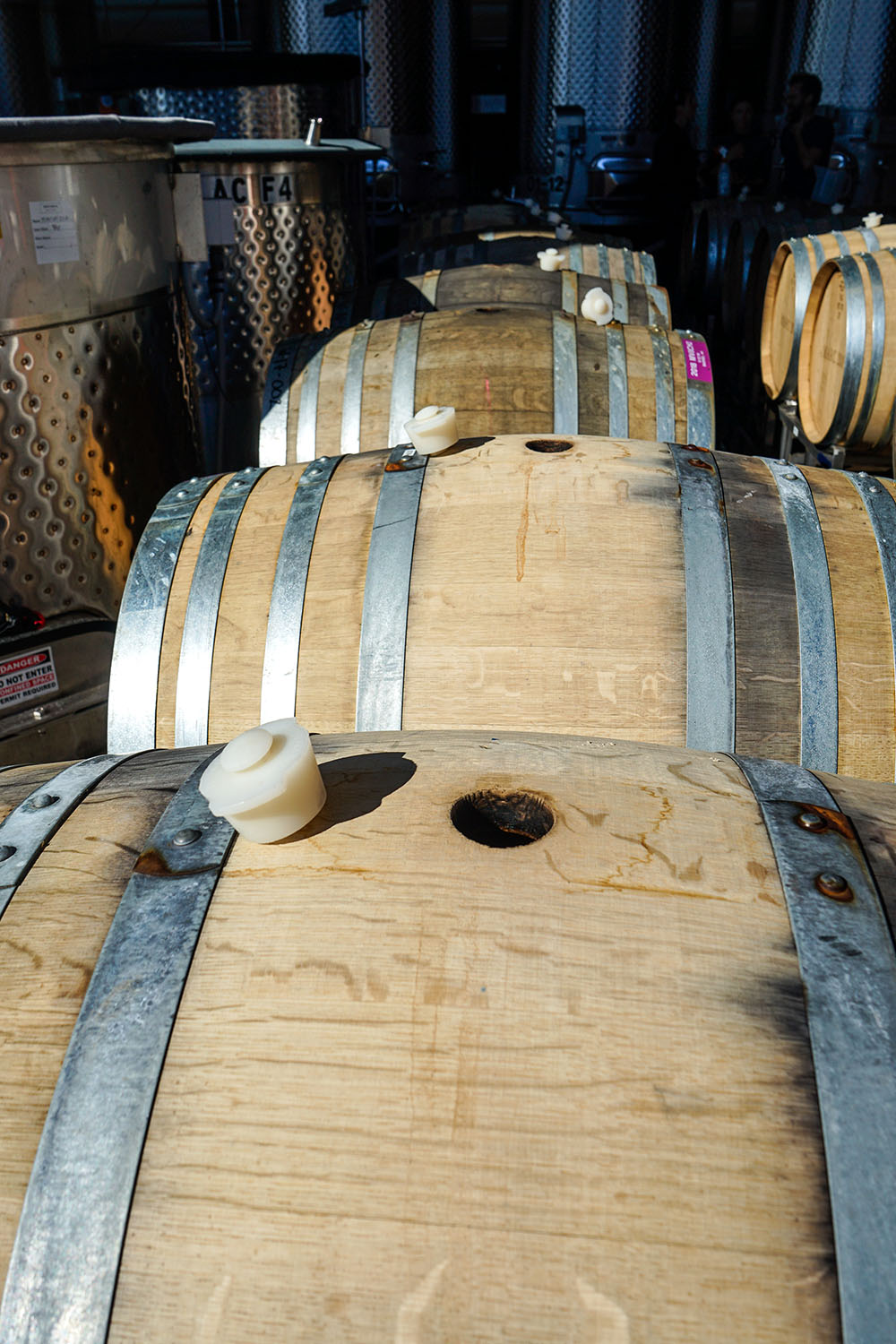 Rows of wooden barrels with metal bands stored in a dimly lit winery or brewery with stainless steel tanks in the background.