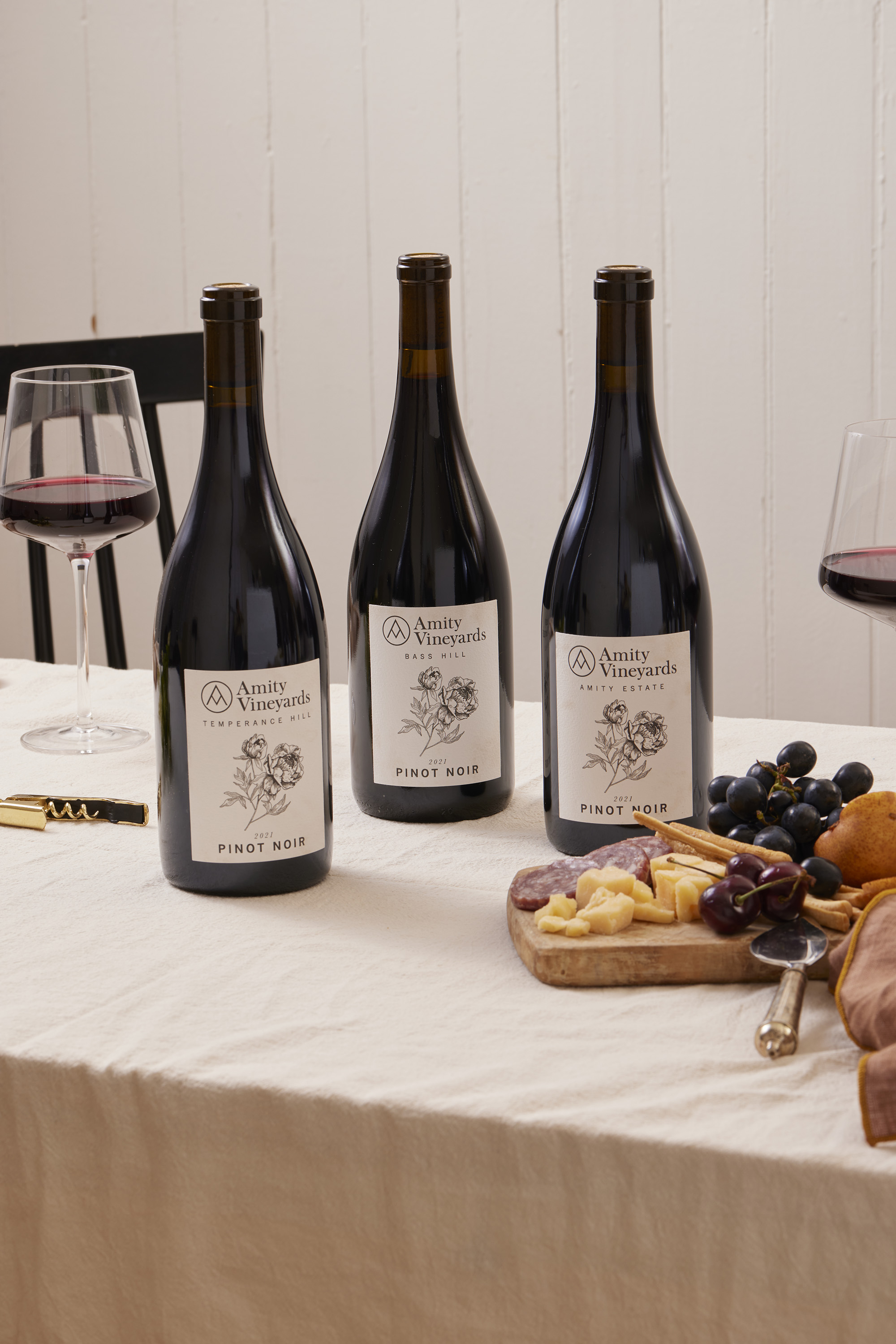 Bottle of 2018 Amity Vineyards Pinot Noir with two glasses of red wine, plums, cinnamon sticks, and pieces of dark chocolate on a light wooden surface.
