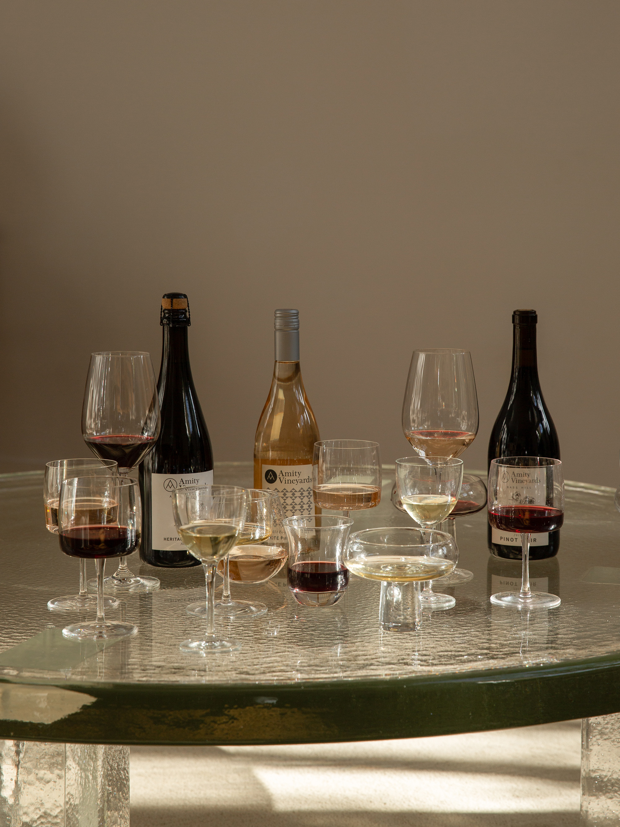 Table set outdoors with three bottles of Amity Vineyards white wine, four wine glasses with white wine, a bowl of red grapes, peaches, cherries, and green leaves.