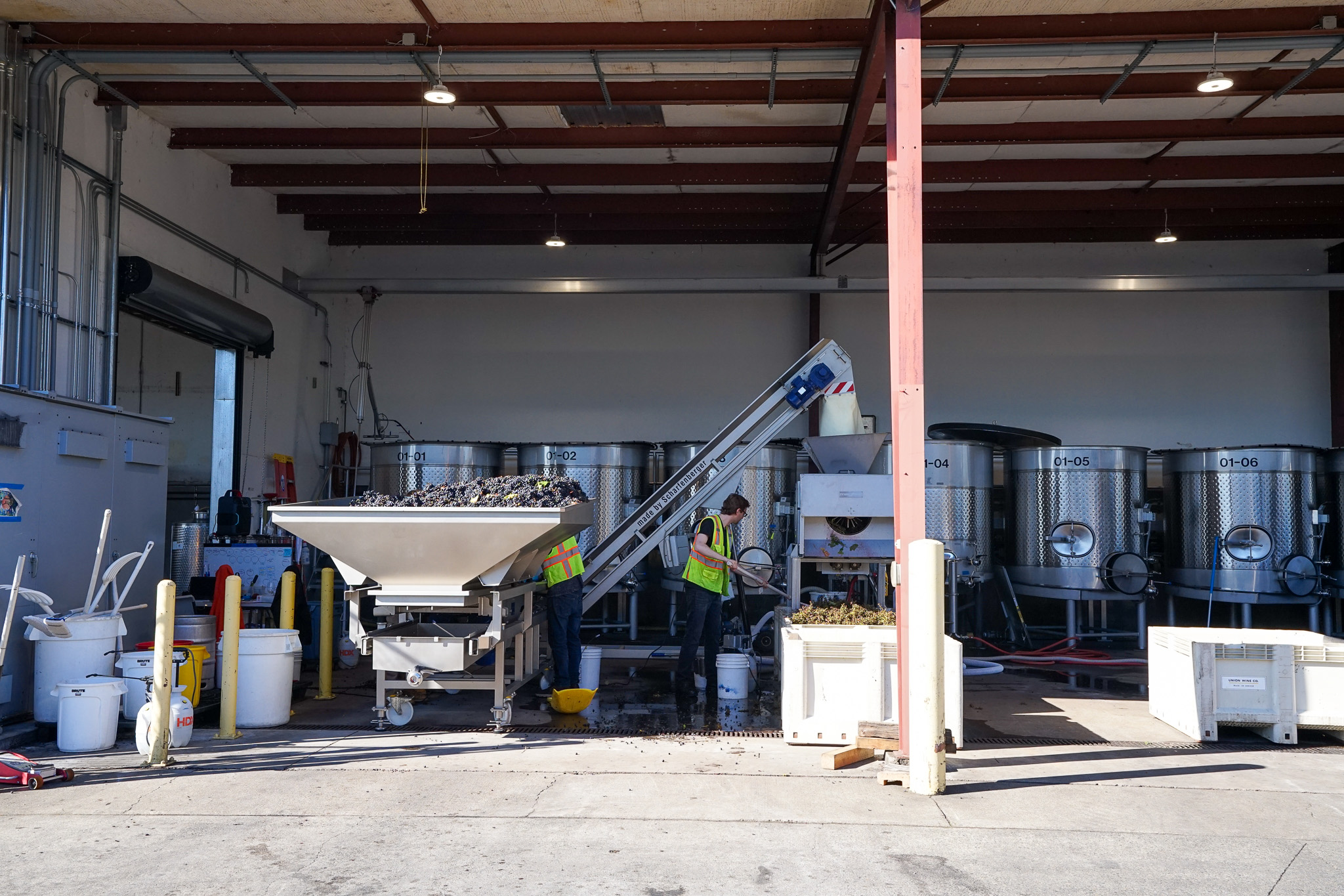 Four people outside an industrial building with large metal tanks inside, one person walking on the right, three standing near entrance.
