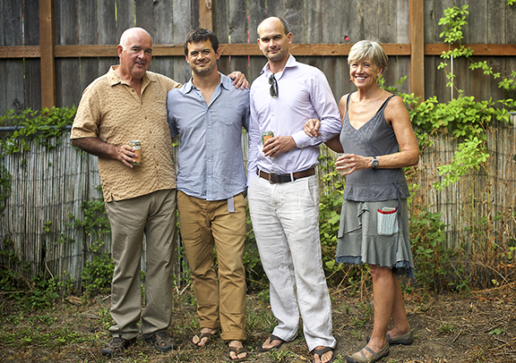 Four adults standing side by side outdoors near a wooden fence, smiling, two holding cans and one holding a glass.