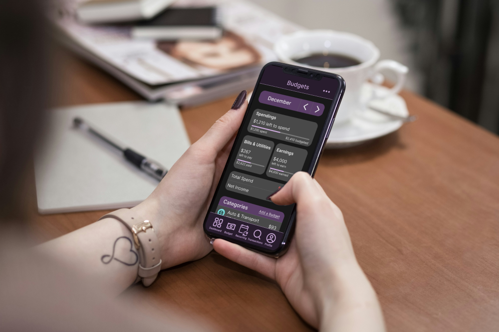 a person wearing a beige watch on the left wrist holding a phone with the budget page opened. she is sitting at a table scrolling on the phone using her right thumb. in the background there is a cup of coffee on a saucer and a spoon, a notepad and pen, and a stack of magazines.