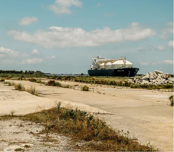 Large cargo ship with spherical tanks docked near rocky shore under a partly cloudy sky.