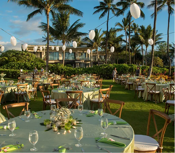 Outdoor wedding reception setup with round tables, green linens, palm trees, and hanging white lanterns at sunset.