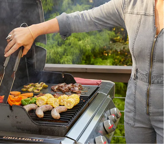 Person grilling sausages, corn, bell peppers, mushrooms, and steak on an outdoor gas grill.