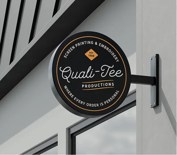 Round black sign for Quali-Tee Productions, a screen printing and embroidery shop, mounted on a gray wall.