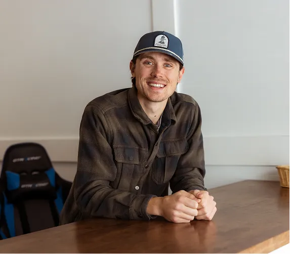 Smiling man in a dark plaid shirt and baseball cap sitting at a wooden table in a light room.