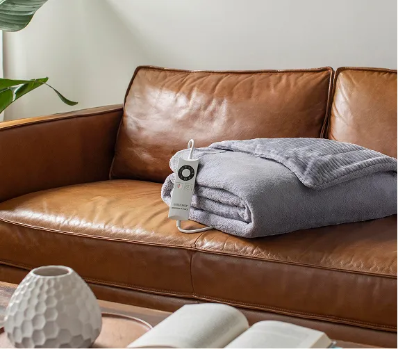 Folded gray electric blanket with remote control on a brown leather couch in a living room.