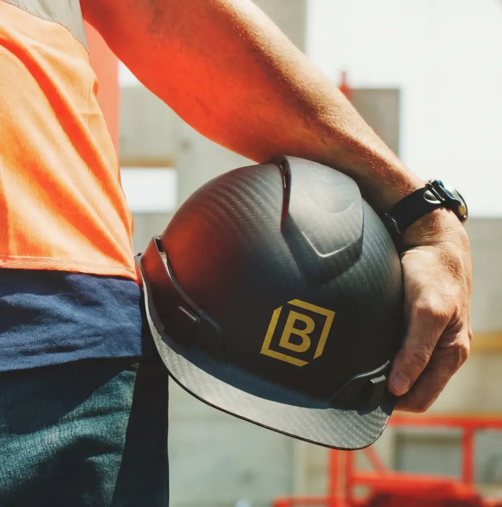 Person wearing orange and blue holds a black hard hat with a yellow B logo at a construction site.