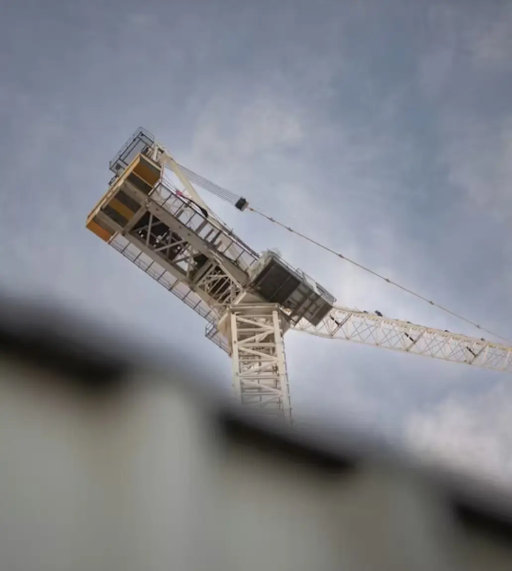 White construction crane against a cloudy sky with a blurred building roof in the foreground.