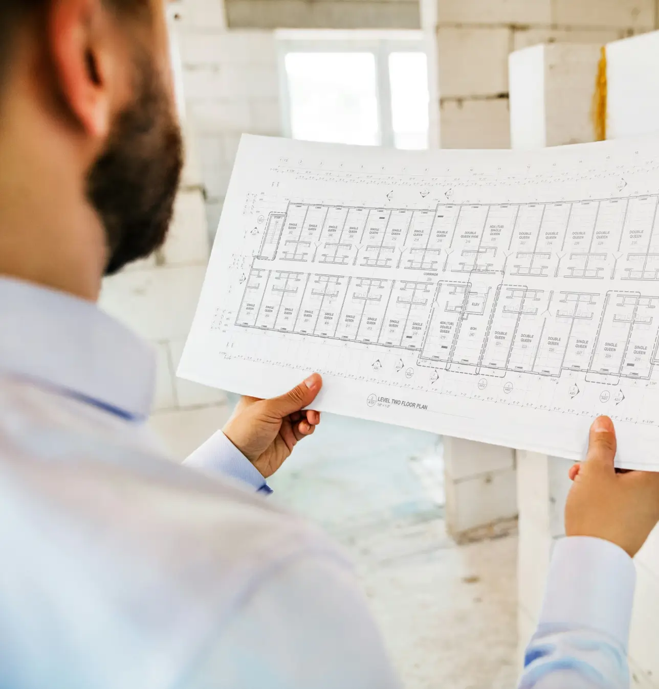 Person in a light blue shirt holding and examining a detailed architectural floor plan inside a construction site.