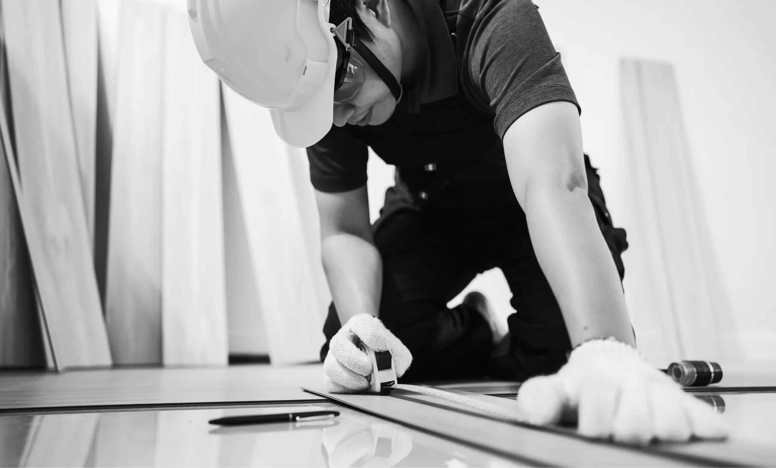 Construction worker wearing a hard hat and gloves measuring wood with a tape measure on the floor.