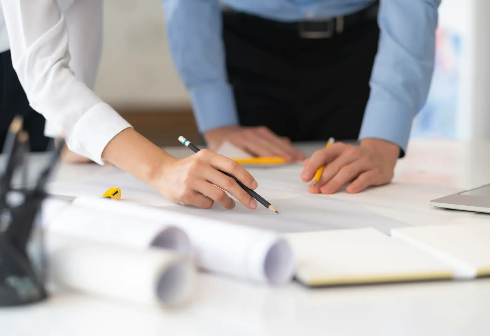 Two people reviewing and marking architectural blueprints on a table with pencils and rolled-up plans nearby.