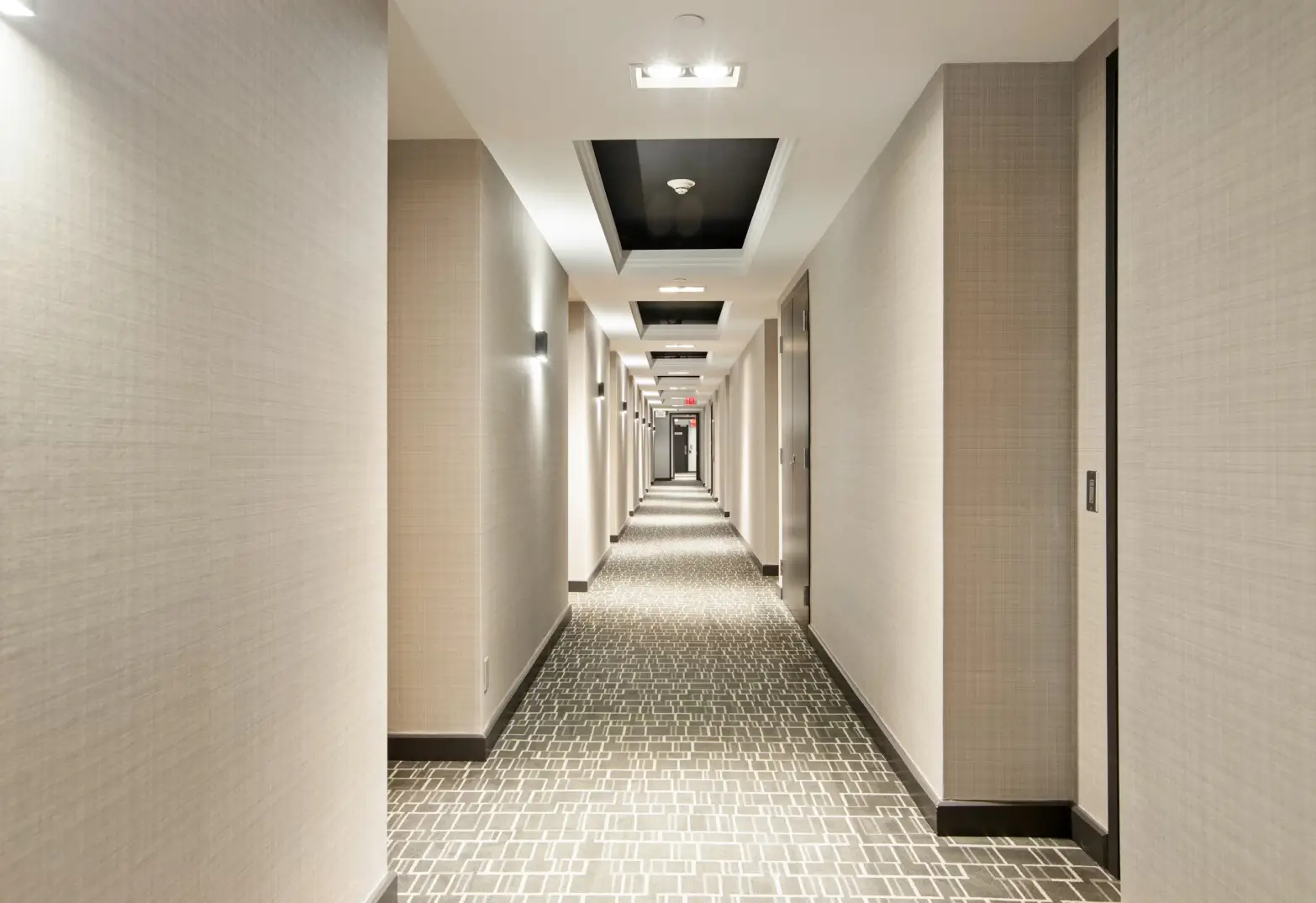 Long hotel hallway with beige walls, patterned carpet, recessed ceiling lights, and closed room doors on the right side.