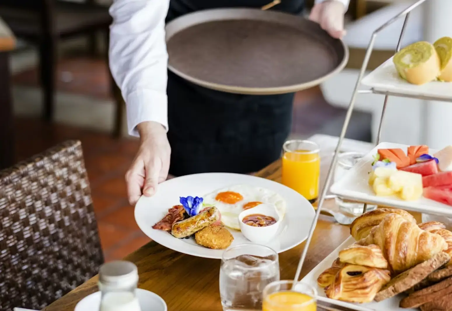Waiter placing a plate with two sunny-side-up eggs, sausage, bacon, hash brown, and a small bowl of baked beans on a breakfast table with juice, water, pastries, and fruit.