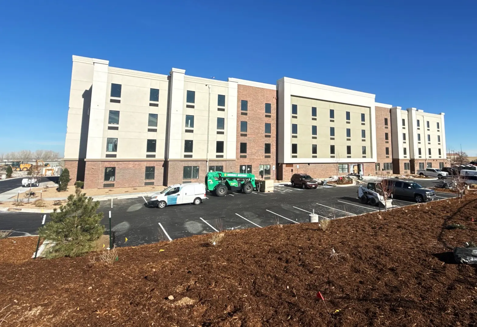 Four-story hotel building under construction with a parking lot and construction vehicles in front against a clear blue sky.